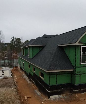 Roof installation on a new home under construction with dark shingles and green exterior wrapping